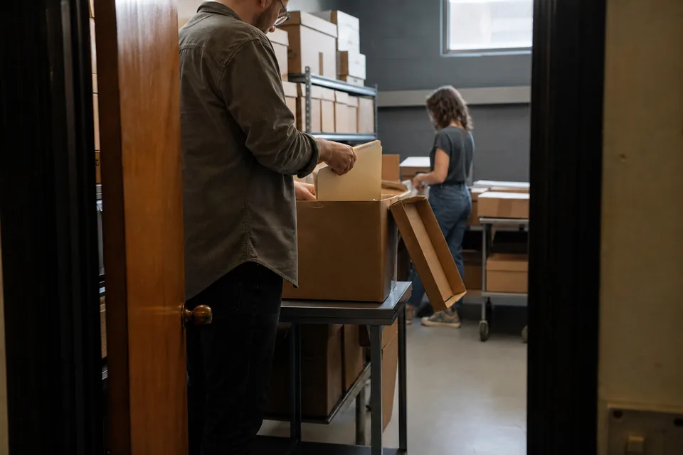 Hands lifting a folder out of an open cardboard archive box on a table, more boxes stacked behind