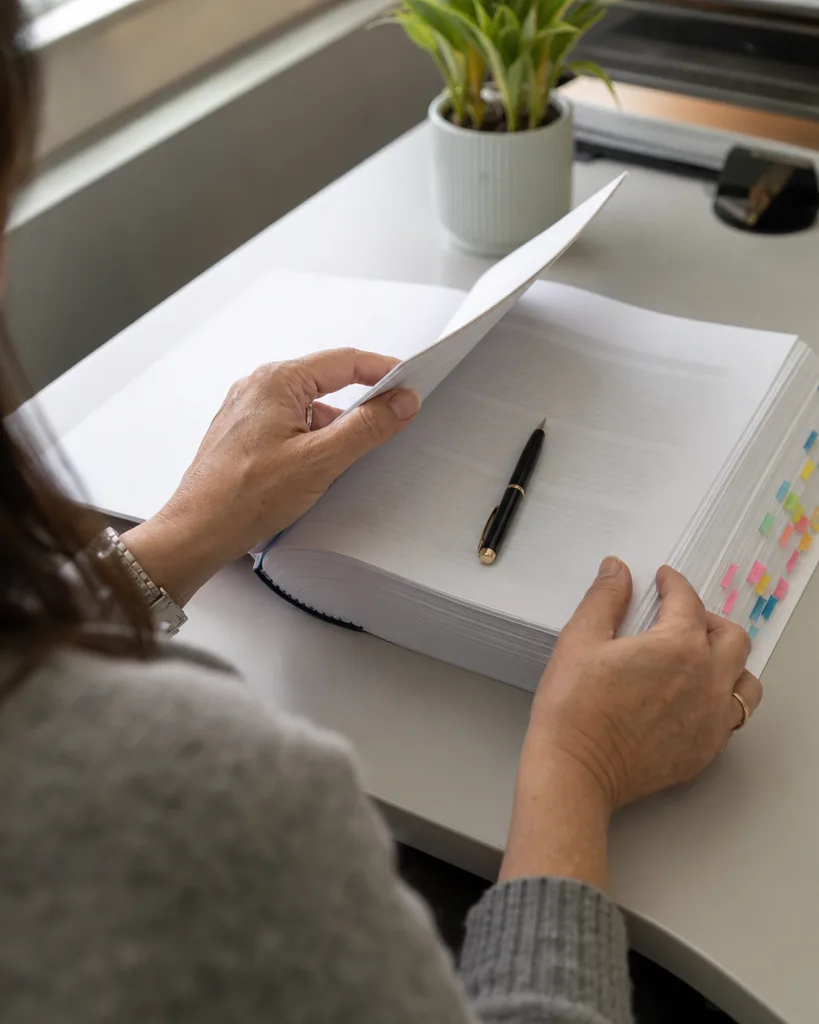 Hands of a paralegal turning a page of a printed contract marked with coloured sticky tabs on a desk by a window