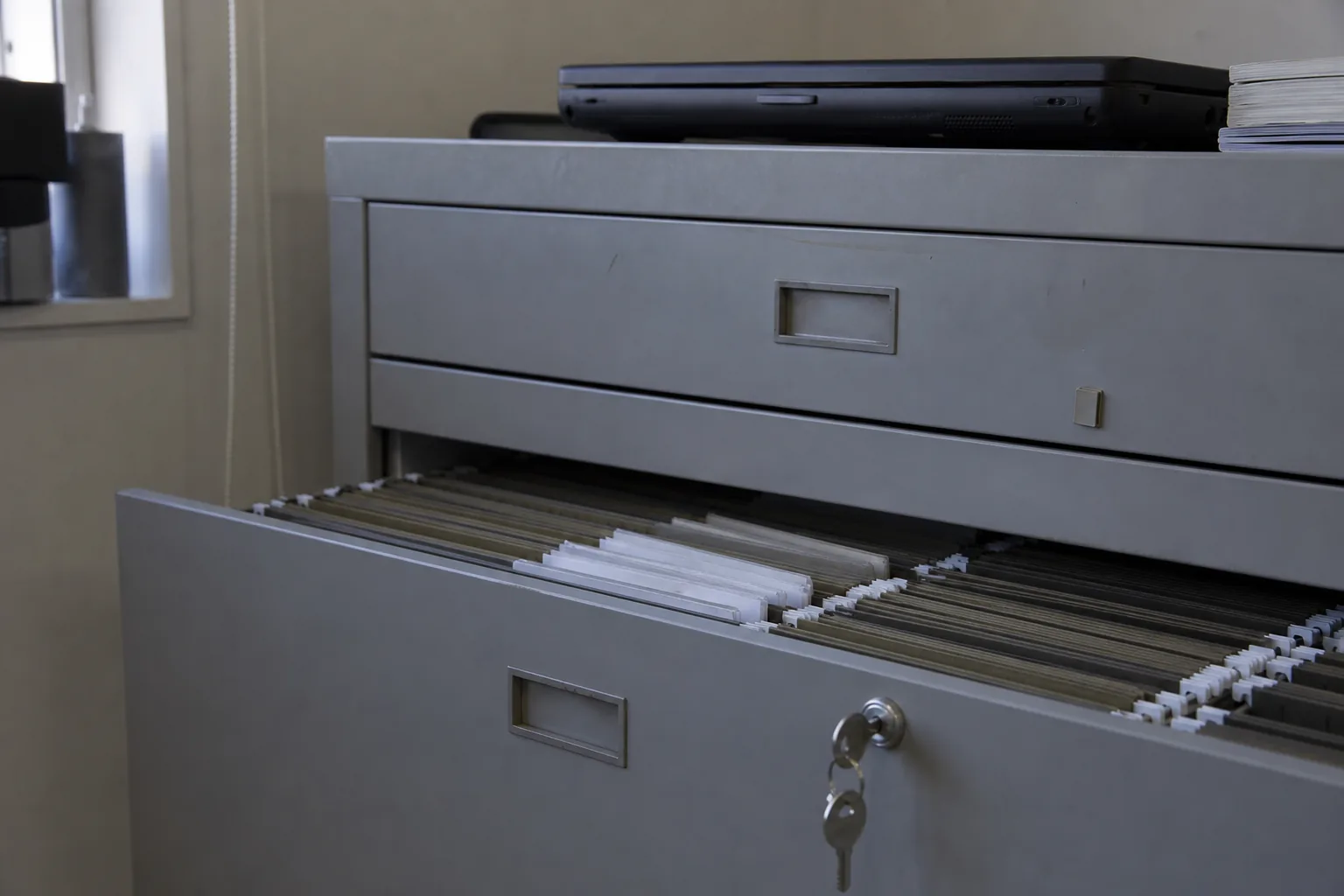 A steel filing cabinet drawer half open with hanging folders, a key in the lock and a closed laptop on top